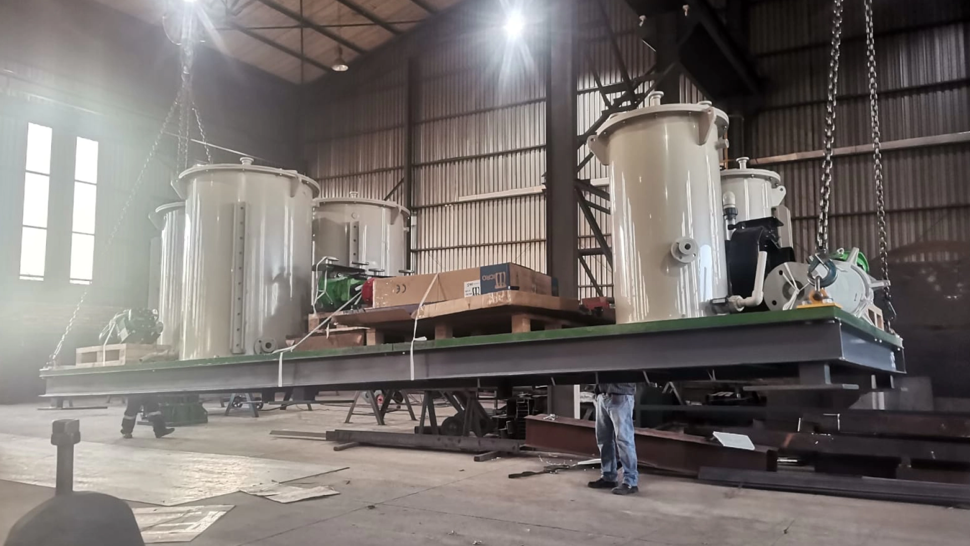 Skid-mounted modular process plant module with tanks and pumps being lifted by crane in a MET63 fabrication workshop.